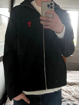 Svart jacka från Ami Paris med röd logga - Snygg svart jacka från Ami Paris med dragkedja och röd hjärtlogga på bröstet. Jackan har en enkel, rak passform och är perfekt för dig som gillar stilrena plagg med en twist. Materialet ser ut att vara bomull eller polyesterblandning.