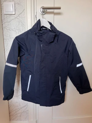 Mörkblå vindjacka med reflexdetaljer, POP  - Snygg mörkblå vindjacka från Polarn O. Pyret med reflexdetaljer på ärmar och rygg. Jackan har hög krage, dragkedja med skyddande vindslå och två fickor framtill. Perfekt för blåsiga dagar och enkel att matcha med andra plagg. (Ingen luva)