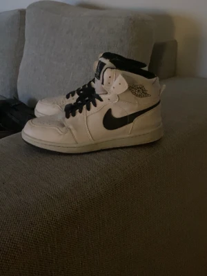 Nike Air Jordan 1 High vita/svarta - Säljer ett par klassiska Nike Air Jordan 1 High sneakers i vitt med svarta detaljer och svarta skosnören. Skorna har den ikoniska swoosh-loggan på sidan och Air Jordan-loggan vid ankeln. Tillverkade i läder med perforerad tå för extra ventilation.