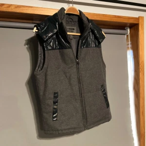 Väst med luva - Snygg och riktigt unik grå väst med svarta quiltade detaljer på axlarna och huva. Västen har dragkedja framtill och praktiska fickor med svarta knappar. Perfekt för lager-på-lager under kyliga dagar. //// priset är inte hugget i sten 