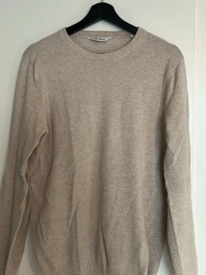 Beige stickad tröja från John henric - En stilren beige stickad tröja från John Henric med rund halsringning och diskret strukturmönster. Tröjan har långa ärmar och ribbade muddar vid ärmslut och nederkant. Perfekt för lager på lager och enkel att matcha.
