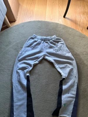 Grå mjukisbyxor Gallery Dept. - Säljer ett par grå mjukisbyxor från Gallery Dept. med svart tryck på vänster sida. Byxorna har elastisk midja, svarta paneler längs benen och färgstänk som ger en unik look. Tillverkade i mjukt sweatshirtmaterial för maximal komfort.