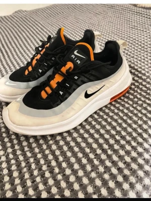 Nike Air Max 98 svart/vit/orange - Nike Air Max 98 sneakers i svart, vitt och orange med synlig Air-sula. Ovandelen är i mesh och syntetmaterial, med tydliga orange detaljer på plösen och snörningen. Klassisk Nike Swoosh på sidan och AIR-tryck på plösen. Skön dämpning och sportig look.