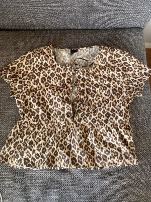 Leopardmönstrad blus med knyt - Snygg blus med leopardmönster och korta ärmar. Blusen har en rund halsringning och knytning framtill samt volang nedtill för en trendig look. Perfekt för dig som vill sticka ut med ett djurmönster. Materialet känns lätt och luftigt.