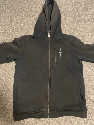 Svart Sail Racing hoodie med dragkedja - Svart hoodie från Sail Racing med hel dragkedja framtill, huva med dragsko och en bröstficka med dragkedja. Mjuk insida och två sidofickor. Perfekt för en sportig och avslappnad stil.