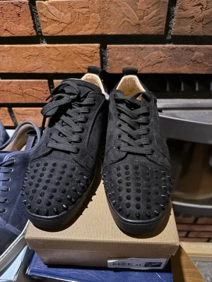 Svarta sneakers med nitar från Louboutin - Svarta sneakers i mocka från Christian Louboutin med svarta nitar på tån och klassisk röd sula. Skorna har svarta skosnören och en låg siluett. Perfekta för dig som vill sticka ut med en edgy och lyxig stil. De har lite svart märke på sulan som behöver special rengöring eller nått åt den stilen