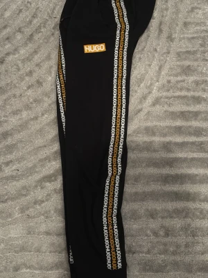 Svarta mjukisbyxor från HUGO - Svarta mjukisbyxor från HUGO med logga i gult på vänster ben och vita/gula loggband längs båda sidorna. Byxorna har dragsko i midjan och smal passform med muddar vid bensluten. Perfekta för en avslappnad och trendig stil.