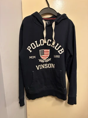 Mörkblå hoodie Polo Club Vinson - Mörkblå hoodie från Polo Club Vinson med vit brodyrtext och amerikansk flagga på bröstet. Klassisk känguruficka framtill, vita dragsnören i huvan och ribbade muddar. Perfekt för en avslappnad och sportig stil.