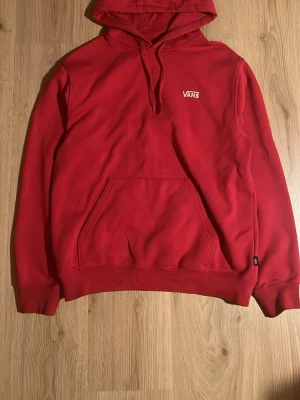 Röd hoodie från Vans med logga - Röd hoodie från Vans med vit broderad logga på bröstet. Klassisk modell med huva, dragsnören och stor magficka framtill. Mjuk insida och ribbade muddar vid ärmslut och nederkant. Perfekt för en avslappnad streetwear-look.