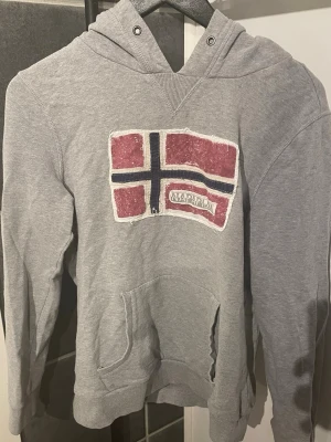 Grå hoodie från Napapijri - Säljer en grå hoodie från Napapijri med stor norsk flagga i rött, vitt och blått på bröstet. Hoodien har huva med dragsko och en stor magficka framtill. Perfekt för dig som gillar streetwear och vill ha en skön och stilren look.
