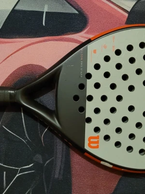 Wilson Pro Staff padelracket - Padelracket från Wilson Pro Staff-serien med svart ram och grå slagyta. Racketen har rund form, orange detaljer och många hål i träffytan för bättre kontroll. Perfekt för dig som gillar padel och vill ha ett snyggt och stabilt racket.