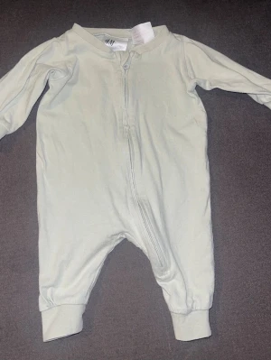 Ljusgrå babyoverall från H&M - Supermjuk och enkel ljusgrå babyoverall från H&M med dragkedja framtill. Perfekt för mysiga stunder hemma eller som pyjamas. Tillverkad i bomull med långa ärmar och ben samt muddar vid ärmslut och benslut.