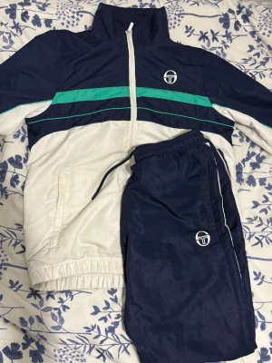 Marinblå och vit träningsdress från Sergio Tacchini - Snygg träningsdress från Sergio Tacchini i marinblått och vitt med gröna detaljer. Jackan har dragkedja, hög krage och logga på bröstet. Byxorna har resår i midjan med snörning och logga på benet. Perfekt för sport eller chill.