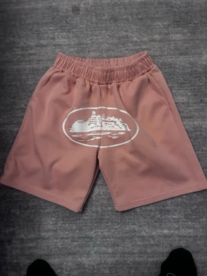 Rosa shorts med tryck och resårmidja - Säljer ett par rosa shorts med vit grafisk print framtill. Shortsen har elastisk midja och en bakficka. Perfekta för chill dagar eller när du vill ha en avslappnad stil. Materialet känns mjukt och bekvämt mot huden. Är storlek m men recommenders till storlek s