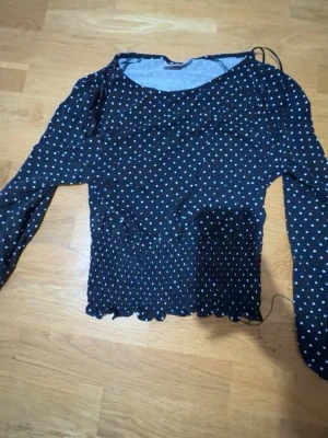 Svart prickig blus Gina Tricot - Snygg svart blus från Gina Tricot med vita prickar. Blusen har lång ärm, smockad midja och vågig kant nertill. Tillverkad i ett tunt och mjukt material som känns skönt mot huden. Perfekt för dig som gillar trendiga mönster och en feminin look.