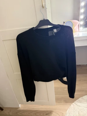 Svart stickad tröja med knytband - Snygg svart stickad tröja med rund halsringning och långa ärmar. Tröjan har ett dekorativt knytband i midjan som ger en trendig siluett. Perfekt att matcha med jeans eller kjol för en cool look.