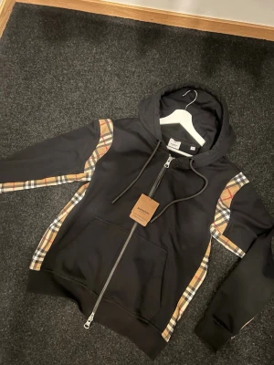 Burberry hoodie - Snygg svart hoodie från Burberry med dragkedja framtill och justerbar huva. Klassisk design med diskreta detaljer och logotyp på snörändarna. Perfekt för en stilren och trendig look. Tillverkad i mjukt material för extra komfort.