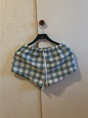 Rutiga shorts med snörning - Säljer ett par rutiga shorts i vitt och ljusgrönt med elastisk midja och snörning framtill. De har en avslappnad passform och är perfekta för varma dagar. Materialet känns som bomull och shortsen har en klassisk, sportig look.