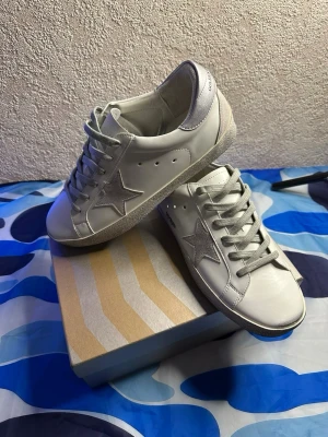 Golden Goose vita sneakers  - Golden Goose sneakers i vitt läder med den ikoniska stjärnan på sidan och grå mockadetaljer vid häl och sula. Skorna har vita snören och en klassisk låg silhuett som ger en clean och trendig look. Perfekta för dig som gillar streetstyle och exklusiva detaljer.
