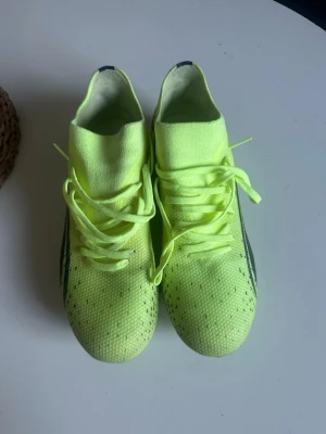 Neongula Puma fotbollsskor - Säljer ett par neongula fotbollsskor från Puma med svart logga på sidan. Skorna har snörning, strumpliknande skaft och dobbar för spel på gräs. Tillverkade i ett lätt syntetmaterial som ger bra bollkontroll och passform. Perfekta för dig som vill sticka ut på planen.