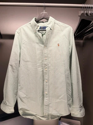  Ralph Lauren - Snygg ljusgrön skjorta från Ralph Lauren med tunna vita ränder och klassisk button-down krage. Skjortan har lång ärm, normal passform och är tillverkad i mjuk bomull. Ikoniska broderade loggan på bröstet och knappar framtill.