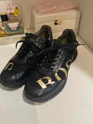 Svarta BOSS sneakers med guldtryck - Stilrena svarta sneakers från BOSS med stora guldiga bokstäver på sidan och BOSS-logga på hälen. Skorna har snörning, svarta sulor och en mix av textil och syntetmaterial. Perfekta för dig som vill ha en clean och lyxig look.