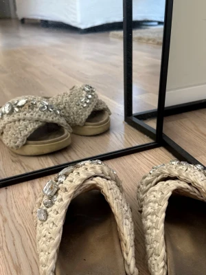 Beige stickade tofflor från Inuikii - Säljer ett par beige stickade tofflor från Inuikii med breda korsade band och stora dekorativa stenar på ovansidan. Tofflorna har en rejäl sula och en mysig, handgjord känsla. Perfekta för dig som gillar unika detaljer och trendig design.