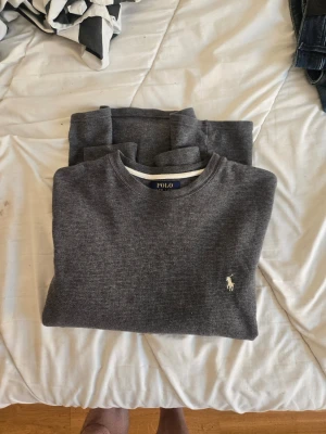 Grå tröja från Polo Ralph Lauren - Mörkgrå långärmad tröja från Polo Ralph Lauren med rund halsringning och den klassiska vita logotypen broderad på bröstet. Tröjan har en enkel och stilren design i mjukt material, perfekt för en clean och avslappnad look.