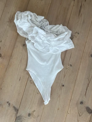 Vit off-shoulder body från Zara - Säljer en vit body från Zara med off-shoulder design och stora volangdetaljer upptill. Materialet är ribbat och stretchigt vilket ger en snygg passform. Perfekt för dig som vill ha något trendigt och unikt i garderoben!