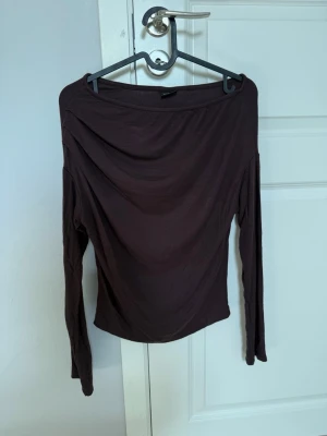 Mörkbrun offshoulder topp - Säljer en mörkbrun offshoulder topp med lång ärm. Toppen har en tight passform och är tillverkad i mjukt, stretchigt material som sitter snyggt på kroppen. Perfekt att styla med jeans eller kjol för en trendig look.