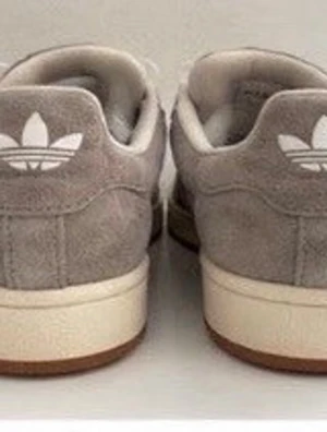 Grå Adidas Gazelle sneakers - Säljer ett par grå Adidas Gazelle sneakers i mocka med klassiska vita ränder på sidan och Adidas-logga på hälen. Skorna har vita snören och en vit sula med brun yttersula. Perfekta för dig som gillar stilrena och tidlösa sneakers.