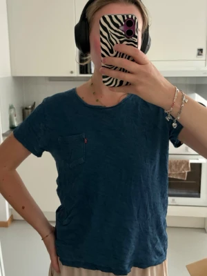Blå Levi's t-shirt med bröstficka - En enkel blå t-shirt från Levi's med rund halsringning och en liten bröstficka med den klassiska röda Levi's-taggen. T-shirten har korta ärmar och är gjord i mjuk bomull.