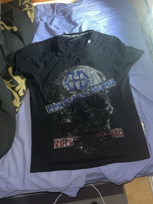 Svart Philipp Plein t-shirt med dödskalle - Säljer en svart t-shirt från Philipp Plein med glittrig dödskalle framtill och detaljer i blått och rött. T-shirten har korta ärmar och rund hals. Materialet är mjuk bomull och modellen är normal passform. Perfekt för dig som gillar statement-plagg!