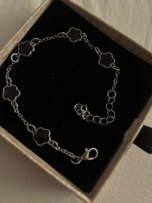 Silverarmband med svarta blommor - Snyggt armband i silverfärg med fem svarta blommor i emalj. Länken är tunn och har justerbar längd med flera öglor.