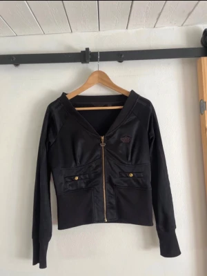 Svart tröja med dragkedja från Adidas - Svart tröja från Adidas med dragkedja framtill och två fickor med guldknappar. Klassisk Adidas-logga på bröstet och långa ärmar. Snygg och sportig design som passar perfekt till streetwear. Materialet känns mjukt och stretchigt.
