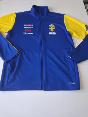 Svenska landslaget träningsjacka blå/gul - Blå träningsjacka från Adidas med gula ärmar. Jackan har hel dragkedja, ståkrage och tryckta sponsorer på framsidan. Materialet är troligtvis polyester och modellen är sportig och bekväm. Varan är i ny skick och i storlek M. Hör av dig vid frågor