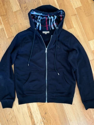 Svart burberry hoodie med dragkedja och mönstrad huva - Säljer en svart hoodie med dragkedja och fickor framtill. Huvan har ett snyggt rutigt foder i rött, vitt och blått som ger en unik touch. Tillverkad i mjukt material med ribbade muddar vid ärmar och nederkant. Perfekt för en chill och stilren look.burberry