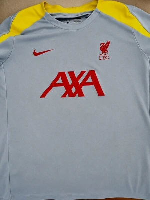 Liverpool tränings-tshirt Nike grå/gul - Snygg Liverpool FC tränings-tshirt från Nike i ljusgrått med gula axelpartier och röda detaljer. Framsidan har Nike-logga, Liverpool-märke och AXA sponsortryck i rött. Tillverkad i lätt och ventilerande funktionsmaterial, perfekt för träning.