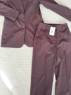 Brun kostym från BIKBOK med kavaj - Snygg brun kostym från BIKBOK med klassisk kavaj och matchande byxor. Kavajen har två knappar, fickor med lock och knappar vid ärmslut. Byxorna har raka ben och pressveck. Perfekt för dig som vill ha en stilren och modern look.