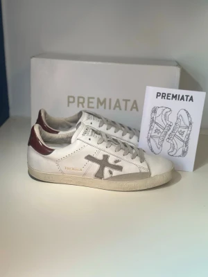 Vita Premiata sneakers med vinröda detaljer - Stilrena vita sneakers från Premiata med grå logga på sidan och vinröda detaljer vid hälen. Skorna har klassisk låg profil, vita snören och sula i gummi. Tillverkade i läder för en exklusiv känsla och passar perfekt till streetwear eller casual outfits.
