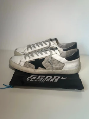 Golden Goose vita sneakers med stjärna - Säljer ett par vita Golden Goose sneakers med grå meshpaneler, svart stjärndetalj på sidan och svart hälparti. Skorna har grå skosnören och klassisk låg siluett. Perfekta för dig som gillar en avslappnad och trendig look.