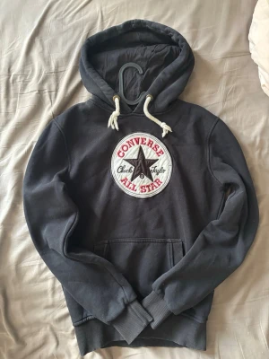Svart Converse All Star hoodie - Säljer en svart hoodie från Converse med stor broderad All Star-logga på bröstet. Hoodien har vita dragsnören, känguruficka och ribbade muddar. Perfekt för dig som gillar streetwear och vill ha en klassisk look med tydlig logga.
