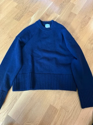 Mörkblå stickad tröja - Mysig mörkblå stickad tröja med rund halsringning och bred ribbstickad kant nedtill samt vid ärmsluten. Tröjan har en avslappnad passform och är perfekt för lager på lager. Passar dig som gillar enkel och stilren design.