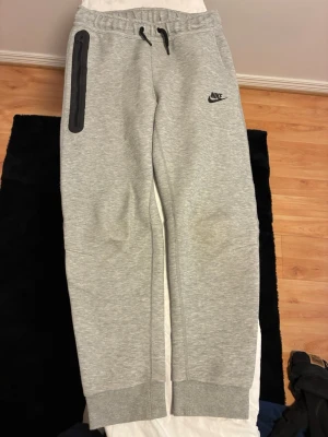 Gråa Nike mjukisbyxor med dragkedja - Snygga grå mjukisbyxor från Nike med svart logga på vänster sida och en cool dragkedjeförsedd ficka på höger lår. Byxorna har resår i midjan med snörning och muddar vid bensluten. Perfekta för chill eller träning, tillverkade i mjukt material.