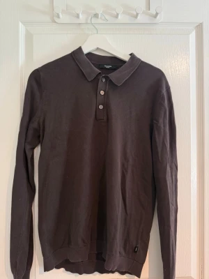 Mörkbrun långärmad pikétröja - Mörkbrun långärmad pikétröja från jack&jones med klassisk krage och tre knappar framtill. Tröjan har ribbade muddar vid ärmslut och nederkant. Perfekt för en stilren och avslappnad look.