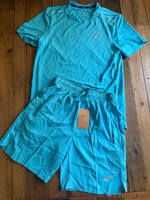 Nike Miller - Snyggt träningsset från Nike i klarblå färg, består av en t-shirt och shorts. Båda plaggen har Nike-logga och är tillverkade i lätt, snabbtorkande Dri-FIT-material som andas. Perfekt för gym, löpning eller annan sport.