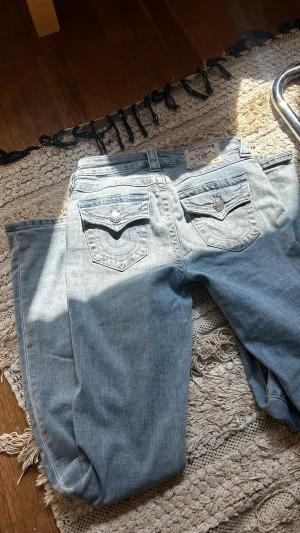 Ljusblå True Religion jeans - Säljer ett par ljusblå jeans från True Religion med ikoniska bakfickor och silverfärgade knappar. Jeansen har utsvängda ben och klassisk femficksdesign. Materialet är denim i bomull och passformen är avslappnad. Perfekta för en cool och avslappnad stil.