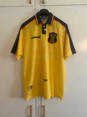 Gul Umbro fotbollströja med klubbmärke - Gul retro fotbollströja från Umbro med svart och röd detalj på axlarna, klubbmärke på bröstet och klassisk krage med knapp. Tillverkad i lätt och ventilerande polyester, perfekt för träning eller match.