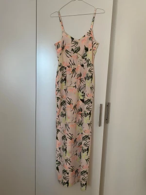 Lång blommig sommarklänning - Säljer en ljus, lång klänning med smala axelband och blommigt mönster i grönt, rosa och gult. Klänningen har v-ringning och slits nertill och är perfekt för varma dagar. Materialet känns lätt och luftigt