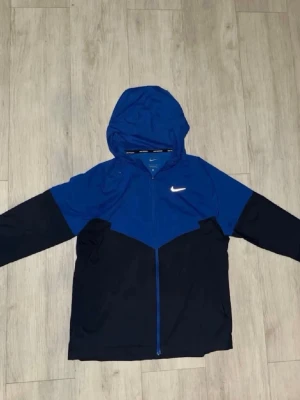Nike blå och svart vindjacka med huva - Snygg vindjacka från Nike i blått och svart med hel dragkedja och huva. Jackan har en sportig look med Nike-logga på bröstet och är perfekt för blåsiga dagar. Lätt material och bekväm passform.